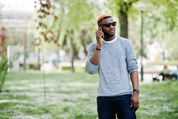 Stylish african american boy on gray sweater and black sunglasses posed on park and speaking on phone Fashionable black guy