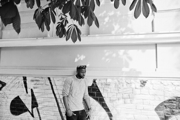 Стильный афроамериканский мальчик в сером свитере и черных солнцезащитных очках позирует на улице Модный темнокожий парень у стены граффити