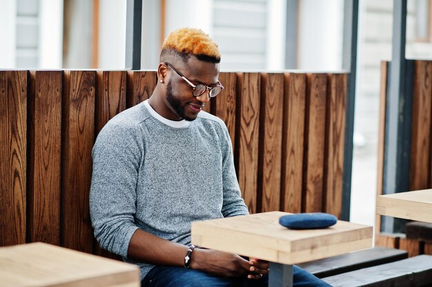 회색 스웨터와 안경을 쓴 세련된 아프리카 소년은 테이블 나무 카페에 앉아 거리에서 포즈를 취했습니다.