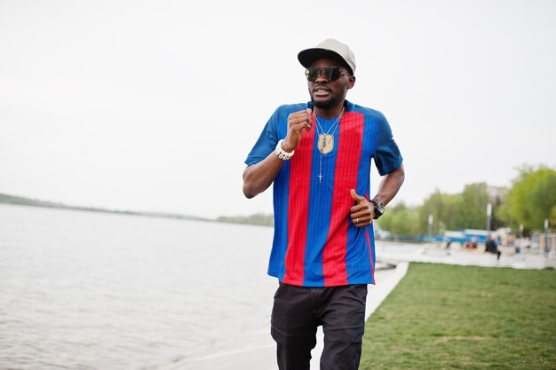 Stylish african american boy running against lake wear at cap football tshirt and sunglasses Black sports man portrait