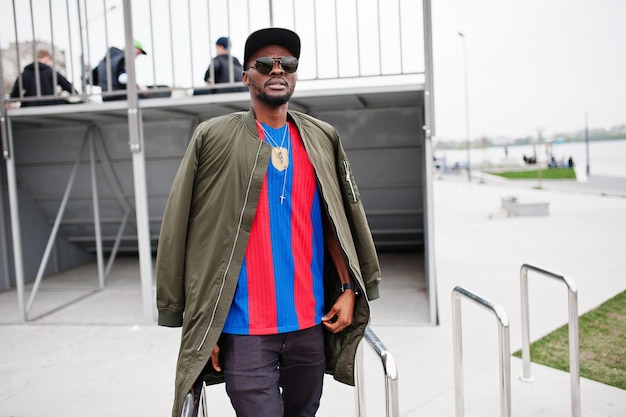 Stylish african american boy wear at cap football tshirt and sunglasses posed on steel railings Black sports man portrait