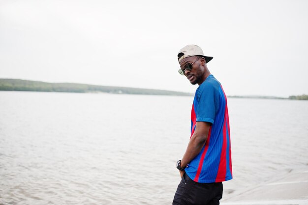 Stylish african american boy wear at cap football tshirt and sunglasses stay against lake beach Black sports man portrait