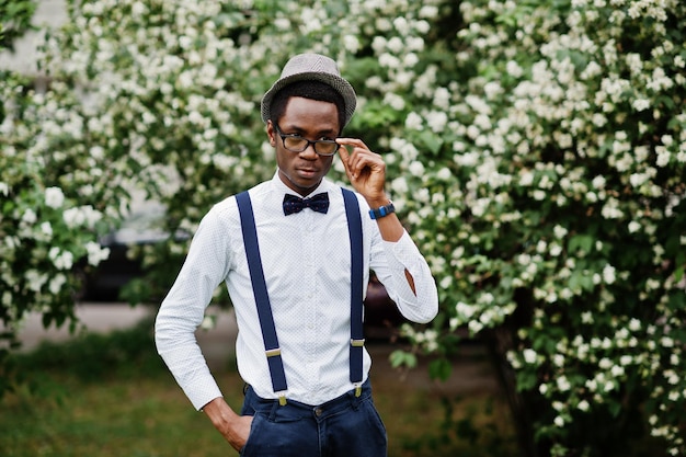 Стильный афро-американский бизнесмен в брюках с подтяжками и рубашке с галстуком-бабочкой, шляпе и очках позирует на открытом воздухе