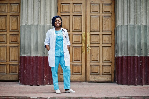 Stylish african american doctor with stethoscope and lab coat posed against door of hospital
