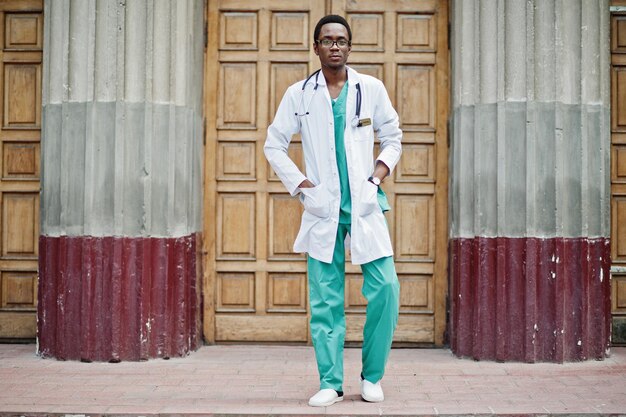 청진기와 실험실 코트를 입은 세련된 아프리카 계 미국인 의사가 병원 문에 포즈를 취했습니다.