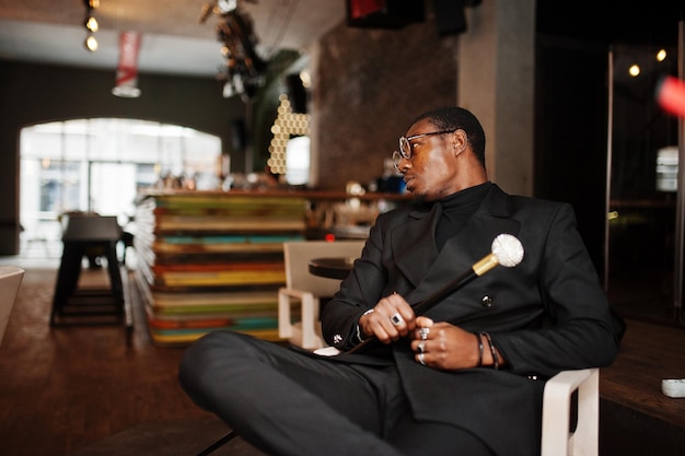 Стильный афро-американский джентльмен в элегантной черной куртке и очках, держащий ретро-трость в качестве тростниковой фляжки или трость с золотым алмазным шариком на ручке Богатый модный афро-мужчина