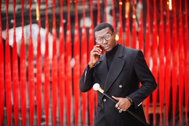 Stylish african american gentleman in elegant black jacket holding retro walking stick as cane flask or tippling cane Rich fashionable afro man against red wall speaking on mobile phone