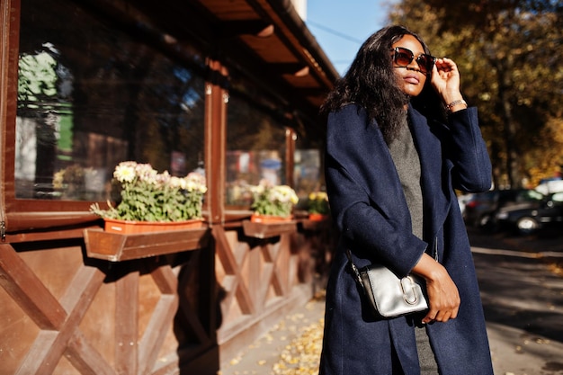 Стильная африканская американка в синем пальто и солнцезащитных очках позирует в солнечный осенний день Африканская модель женщина
