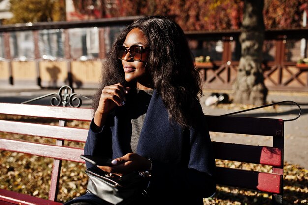 Стильная афроамериканка в синем пальто и солнцезащитных очках позирует в солнечный осенний день на скамейке и пользуется своим мобильным телефоном Африканская модель женщина со смартфоном