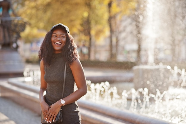 Stylish african american girl in gray tunic crossbody bag and cap posed at sunny autumn day against fountains Africa model woman