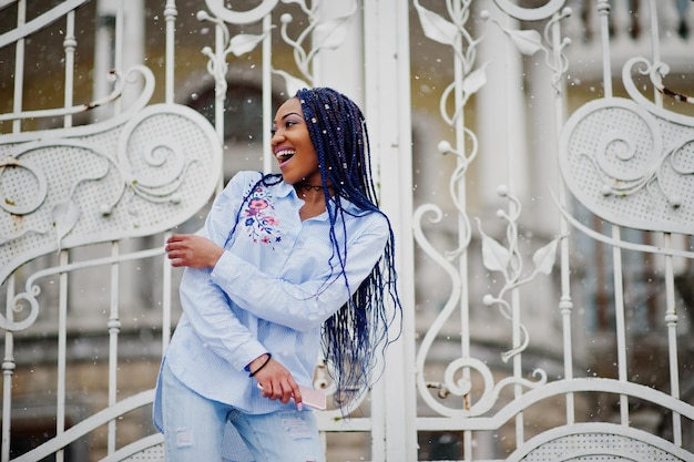 Stylish african american girl with dreads holding mobile phone at hand outdoor against white royal gates at snowy weather