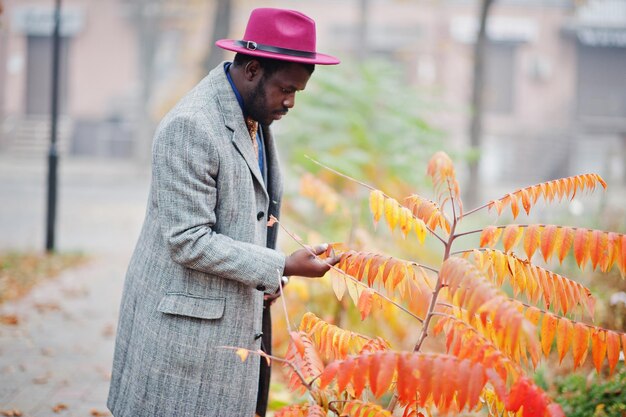 Elegante modello da uomo afroamericano in giacca grigia con cravatta e cappello rosso contro l'atmosfera autunnale di foglie gialle