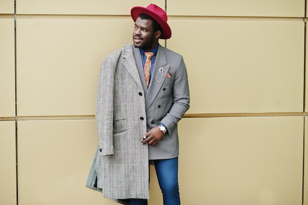 회색 코트 재킷 넥타이와 빨간 모자를 쓴 세련된 아프리카계 미국인 남자 모델이 황금색 배경에 포즈를 취했습니다.