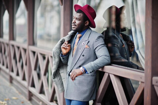 Stylish African American man model in gray coat jacket tie and red hat posed against wooden cafe