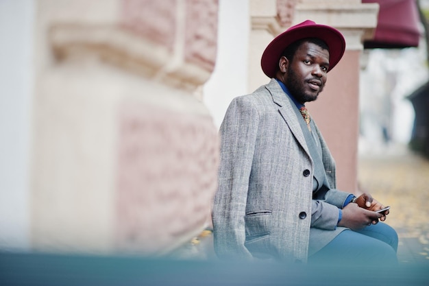 Elegante modello da uomo afroamericano in giacca grigia con cravatta e cappello rosso seduto contro grandi finestre e tenendo il telefono a portata di mano