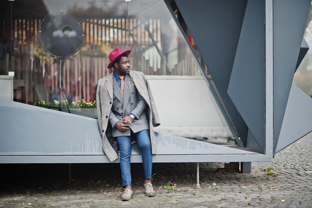 Stylish African American man model in gray coat jacket tie and red hat with mobile phone at hands