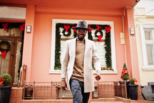 スタイリッシュなアフリカ系アメリカ人の男性は、新年の装飾と花輪で家に対してポーズをとるサングラスとベージュのジャケットと黒の帽子を着用します