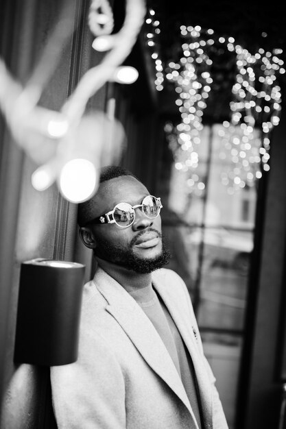 Stylish african american man wear beige jacket with sunglasses pose against house with golden garland