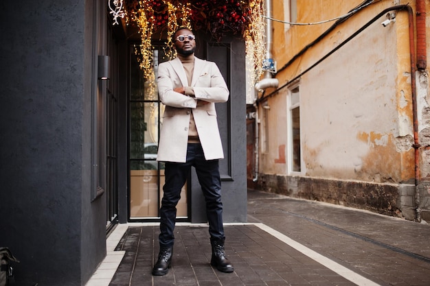 Stylish african american man wear beige jacket with sunglasses pose against house with golden garland