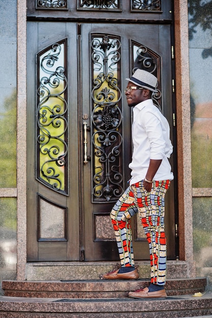 Стильный афроамериканец в белой рубашке и цветных штанах в шляпе и очках позирует на улице Черный модный мальчик-модель