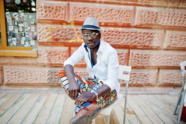 Stylish african american man in white shirt and colored pants with hat and glasses posed outdoor Black fashionable model boy