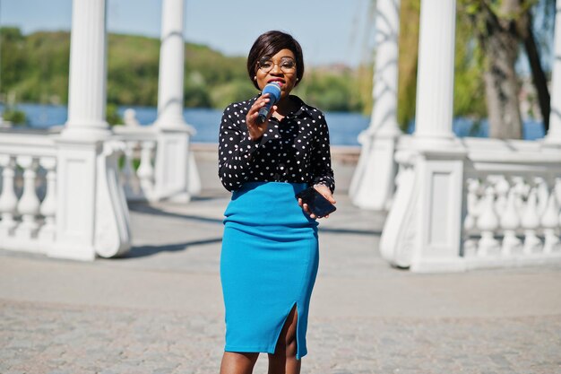 Stylish african american model girl TV presenter with microphone in glasses blue skirt and black blouse posed outdoor against white stone arch with mobile phone