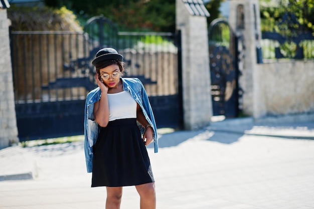 Free photo stylish african american model in glasses hat jeans jacket and black skirt posed outdoor