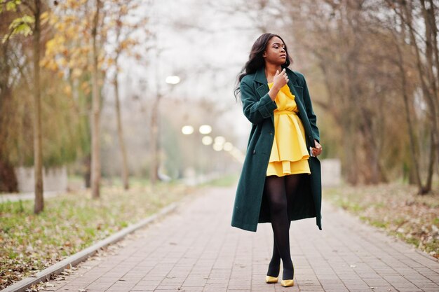 Stylish african american woman at green coat and yellow dress posed against autumn park