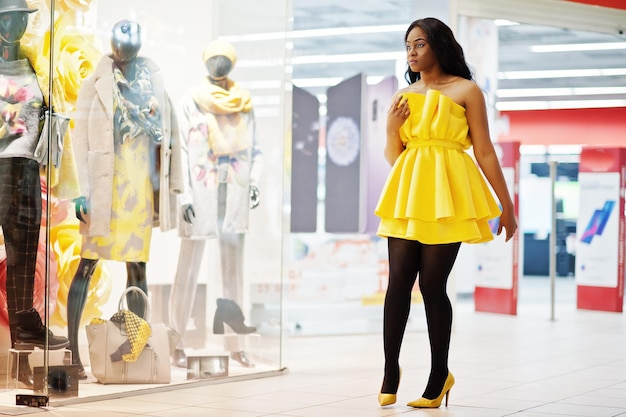 노란색 드레스를 입은 세련된 아프리카계 미국인 여성이 쇼핑몰에서 마네킹이 있는 쇼케이스에 대해 포즈를 취했습니다.