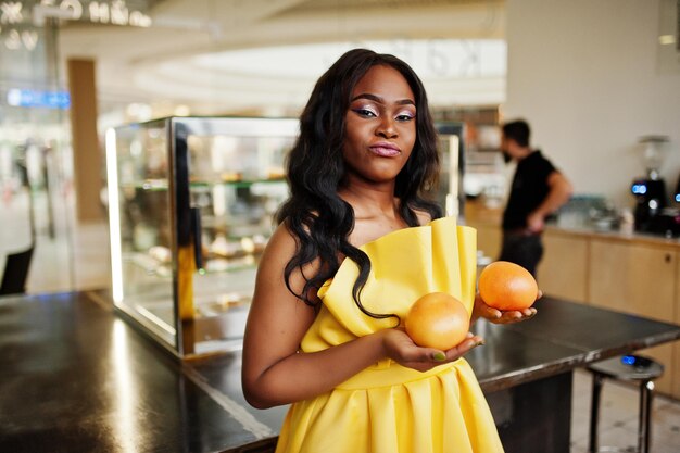 Stylish african american woman at yellow dreess posed at coffee house with two grapefruits on hands
