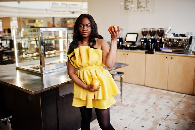 手に2つのグレープフルーツとコーヒーハウスでポーズをとった黄色のドレスでスタイリッシュなアフリカ系アメリカ人の女性
