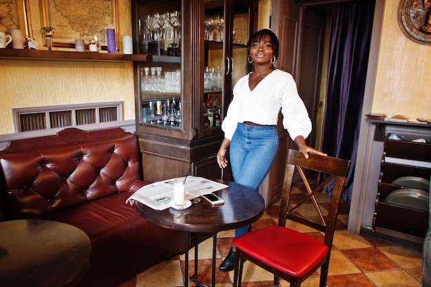 Стильные африканские американки в белой блузке и синих джинсах позируют в кафе с газетой