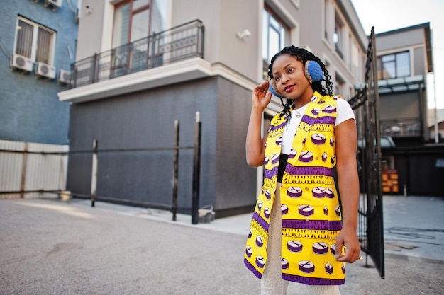 Stylish african american women in yellow jacket posed and earphones on street against modern building