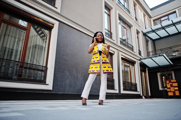 Stylish african american women in yellow jacket posed on street with hot drink in disposable paper cup and mobile phone at hands