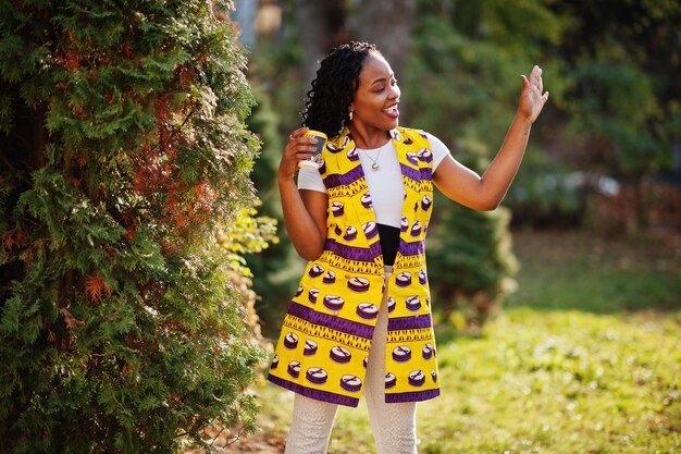 Stylish african american women in yellow jacket posed on street with hot drink in disposable paper cup