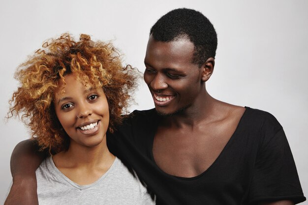 Stylish African couple posing: happy handsome man in black t-shirt hugging his beautiful girlfriend with Afro hairstyle and nose-ring, looking at her with affectionate smile.