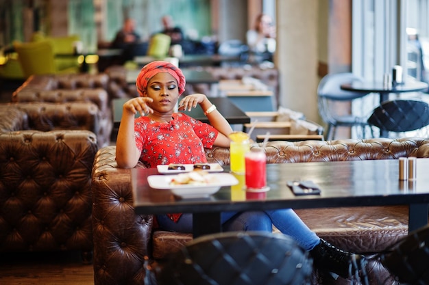 Stylish african woman in red shirt and hat posed indoor cafe and drinking pineapple lemonade