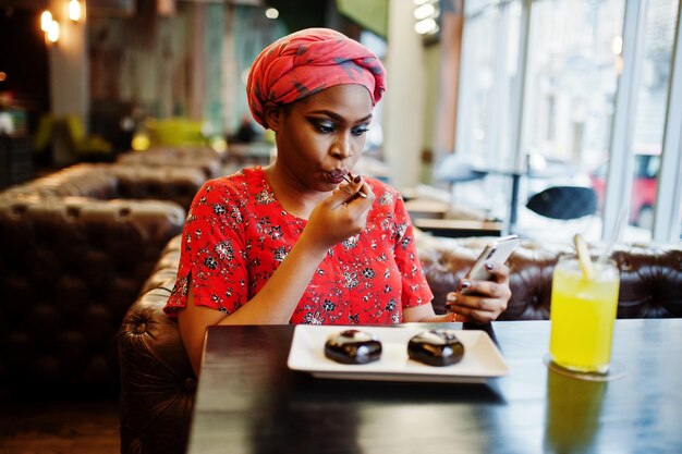 빨간 셔츠와 모자를 쓴 세련된 아프리카 여성이 실내 카페에서 초콜릿 디저트 케이크를 먹고 휴대전화를 보고 있다