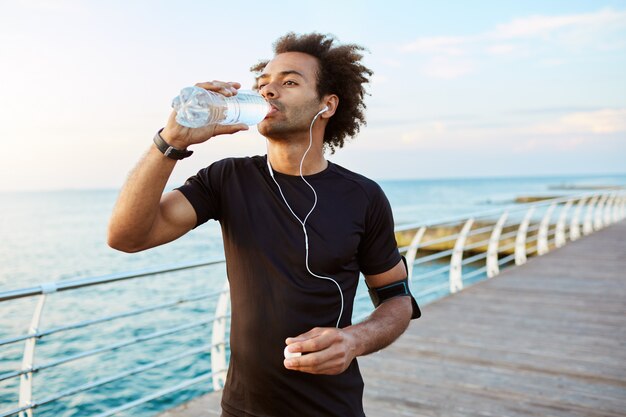 Stylish Afro-American male runner drinking water out of plastic bottle after cardio workout, wearing white earphones. Sportsman in black sportswear hydrating during outdoor training.