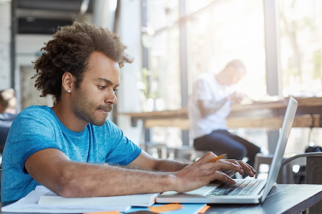 Stylish Afro American student keyboarding on laptop computer while sitting at cafe table with textbooks, working on homework, having focused concentrated look. People, modern technology and education