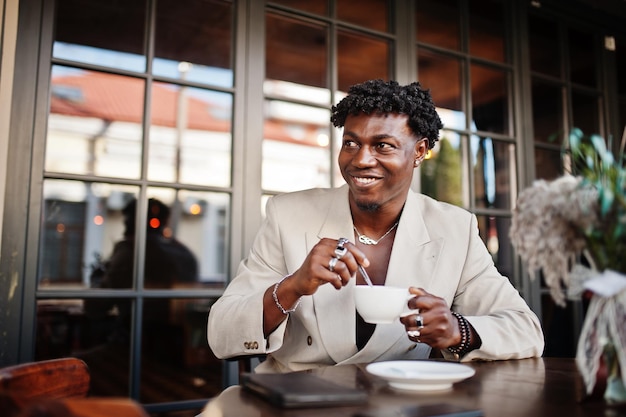 コーヒーとカフェに座っているベージュの古い学校のスーツのスタイリッシュなアフロ男裸の胴体にカジュアルなジャケットでファッショナブルな若いアフリカ人男性