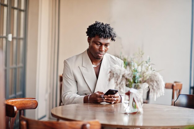 携帯電話でカフェに座っているベージュの古い学校のスーツのスタイリッシュなアフロ男裸の胴体にカジュアルなジャケットでファッショナブルな若いアフリカの男性