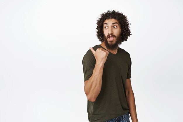 Стильный арабский парень с длинной бородой, указывающий за плечо и смотрящий на рекламную распродажу, стоящую на белом фоне