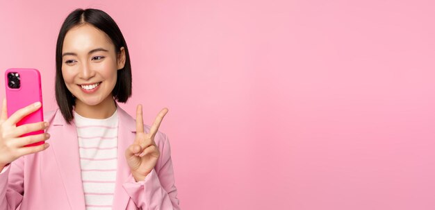 Стильная азиатская деловая женщина в костюме делает селфи в видеочате на смартфоне с приложением для мобильного телефона, позирующим на фоне розовой студии