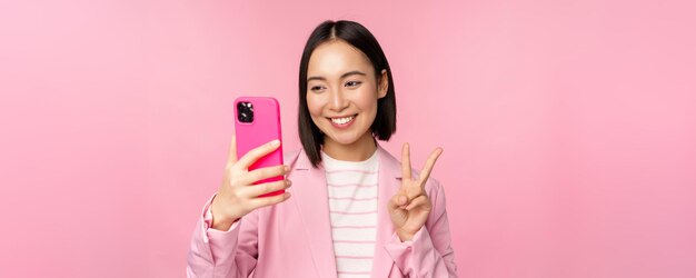 ピンクのスタジオの背景に対してポーズをとって携帯電話アプリとスマートフォンのビデオチャットでselfieを取るスーツのスタイリッシュなアジアの実業家の女の子