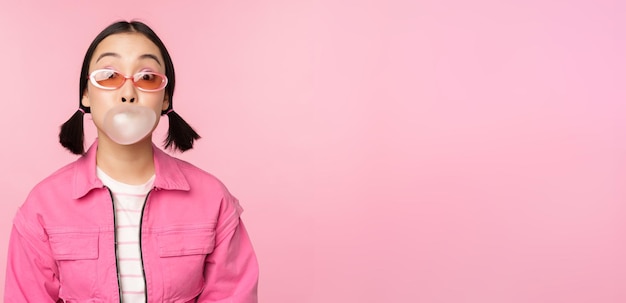 Free photo stylish asian girl blowing bubblegum bubble chewing gum wearing sunglasses posing against pink background