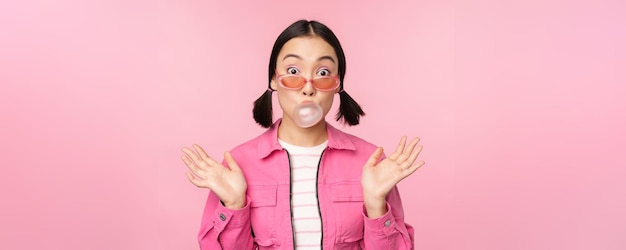 Ragazza asiatica alla moda che soffia gomma da masticare bolla di gomma da masticare indossando occhiali da sole in posa su sfondo rosa