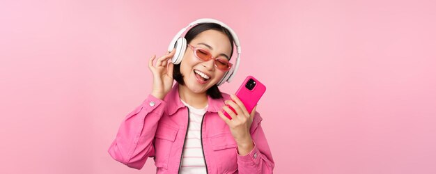 Стильная азиатская девушка танцует со смартфоном, слушая музыку в наушниках в приложении для мобильного телефона, улыбаясь и смеясь, позируя на розовом фоне