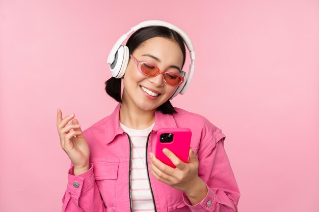 Stylish asian girl dancing with smartphone listening music in headphones on mobile phone app smiling and laughing posing against pink background
