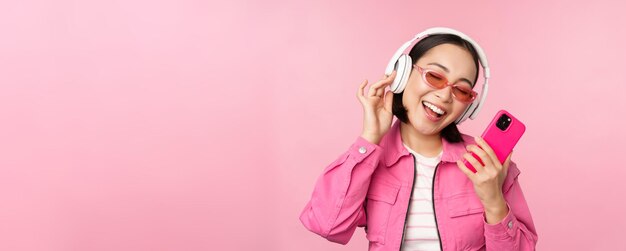 ピンクの背景にポーズをとって笑って笑って携帯電話アプリのヘッドフォンで音楽を聴いてスマートフォンで踊るスタイリッシュなアジアの女の子